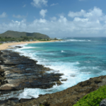 Coastal cliff with rocky shoreline, turquoise ocean, and sandy beach under a blue sky.
